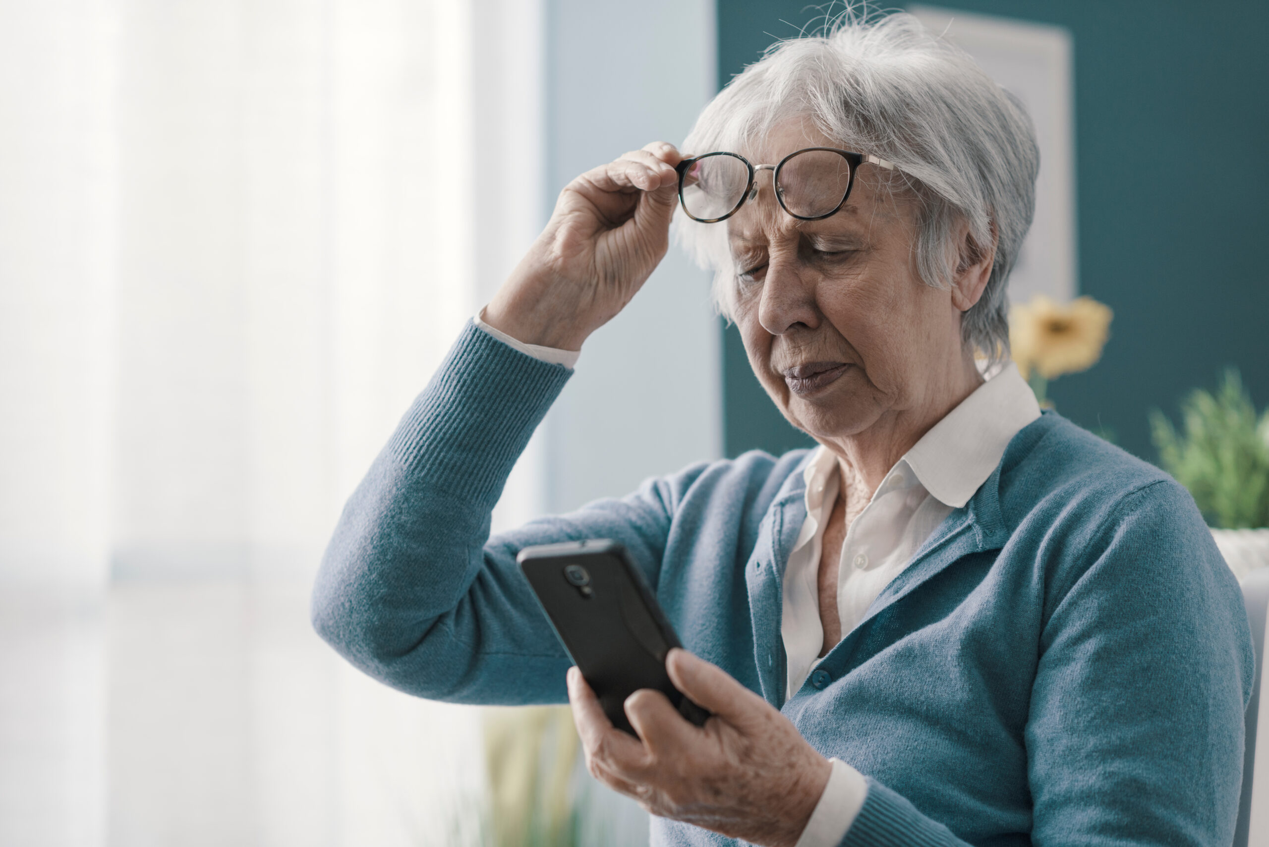 Seniorin hebt Brille an, um auf Smartphone zu fokussieren